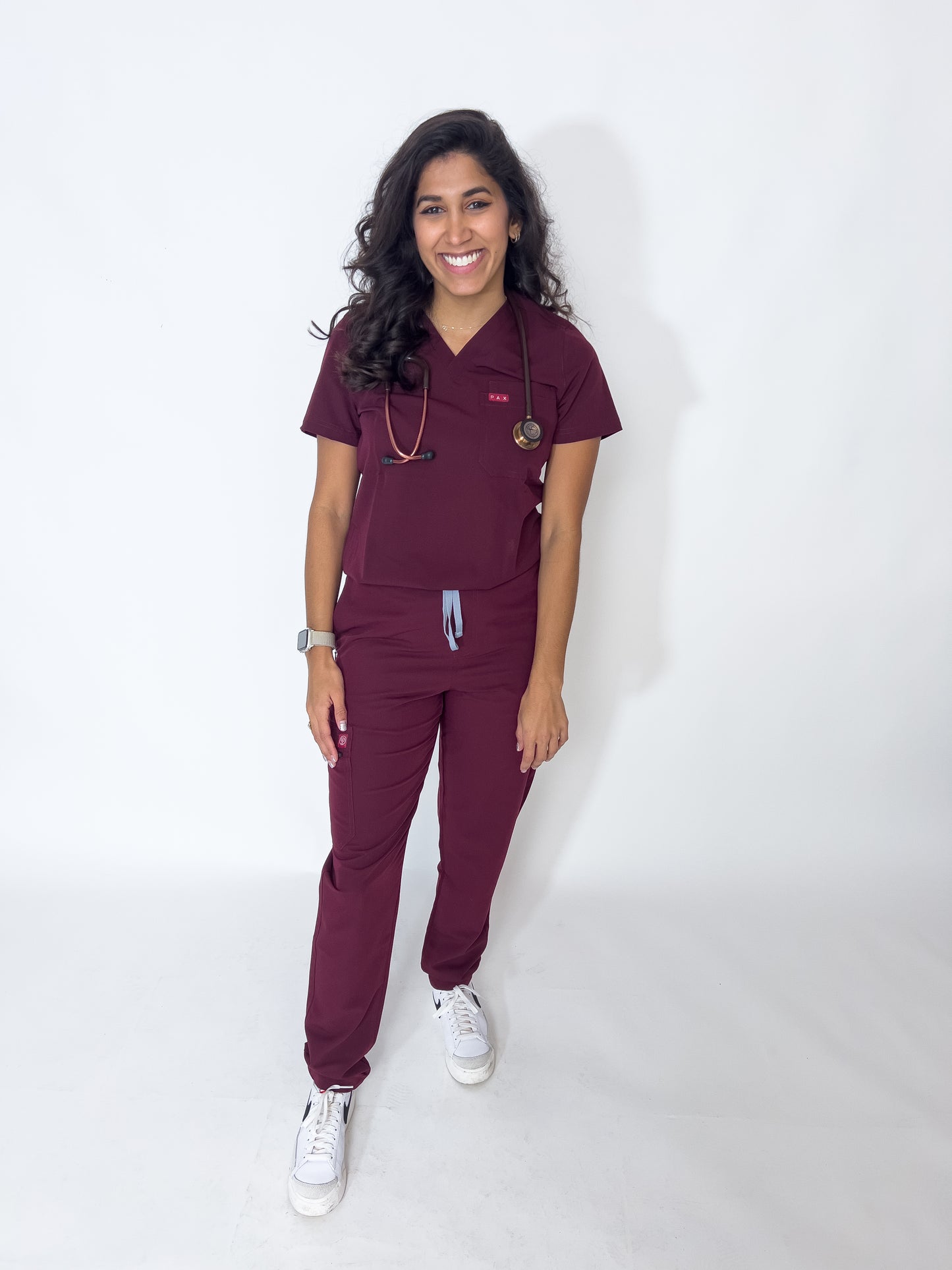 Radiant female doctor in PAX Primus crimson scrubs with stethoscope and white shoes.